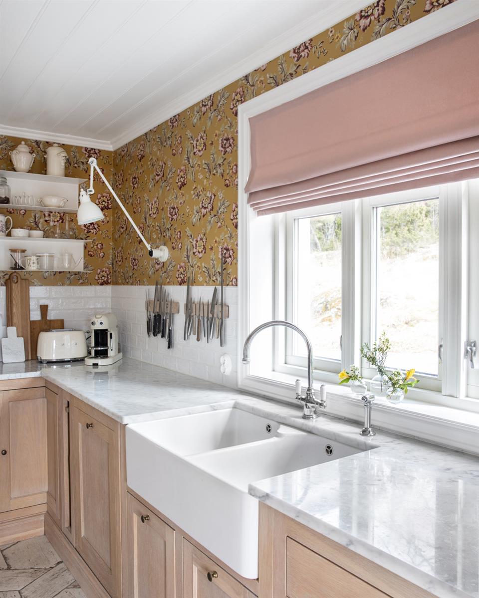 Kjøkken med liftgardin rosa, mønstret tapet og kjøkkeninnredning i tre. Foto.